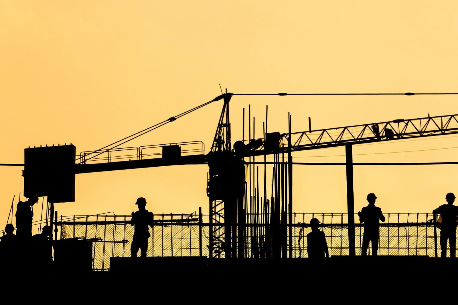 Silhouettes d'ouvriers et d'une grue à tour sur un plancher de coffrage, à contre-jour dans un ciel doré.