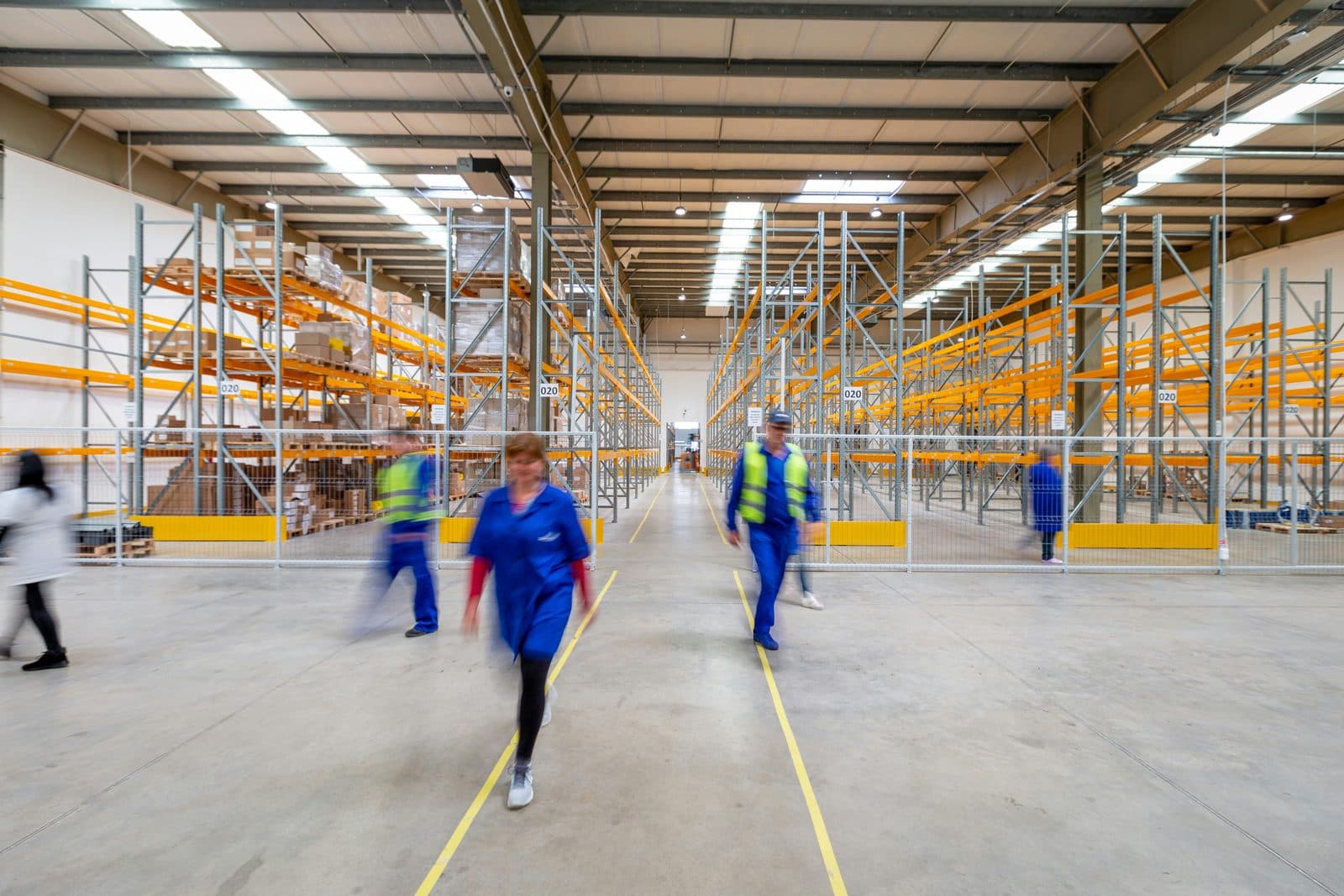 Allée centrale d'un centre de distribution bordée de racks à palettes jaunes, employés en mouvement.