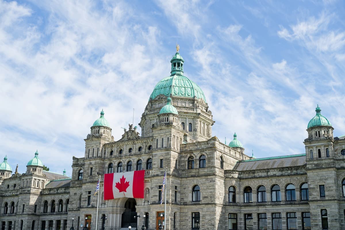 Drapeau canadien flottant devant un édifice du Parlement, sous un ciel dégagé.